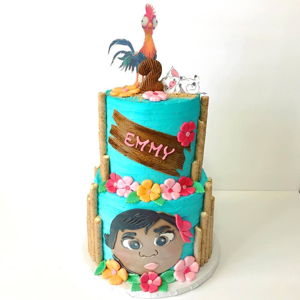 Baby Moana Cake by Hidden Gem Cakes. Two tiered buttercream frosted cake with baby Moana face on the side in fondant. Toppers are Hei Hei and Pua. Pink and orange flowers accent the borders. 